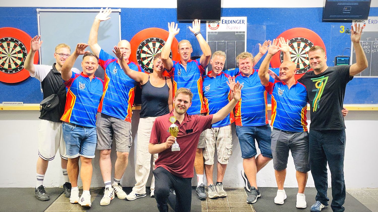 Mitglieder des Dartclub Gamsen 1996 jubeln, Turniersieger Leon Remde kniet mit dem Pokal davor